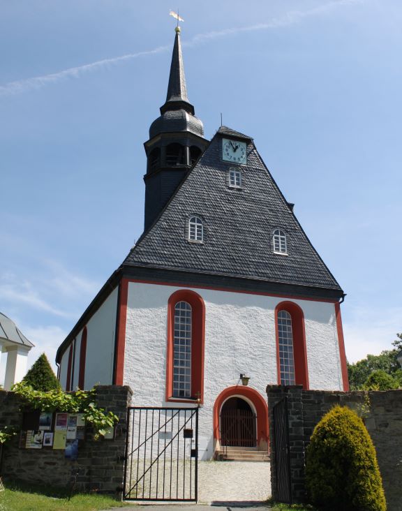 Das Foto zeigt die Trinitatiskirche in Königswalde von der Vorderseite. Zu sehen ist außerdem ein Teil der Friedhofsmauer. Die Tür zum Friedhof steht offen. 